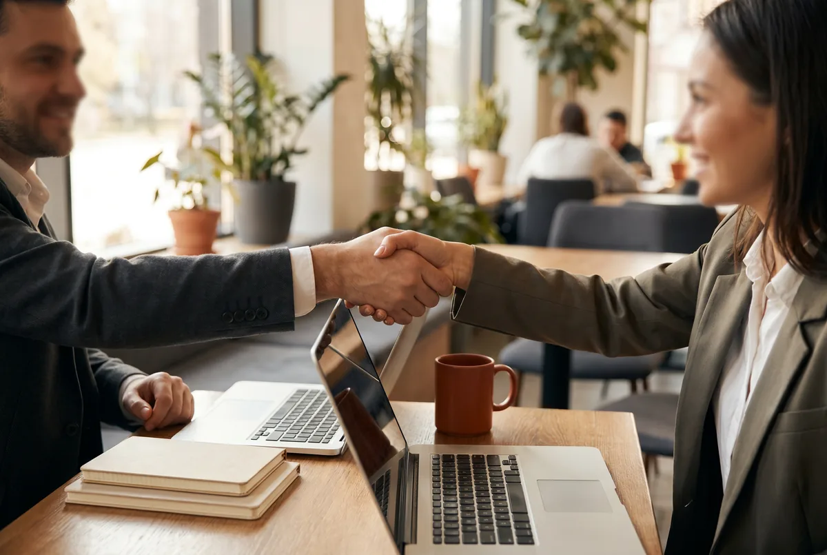Two professionals shaking hands over their laptops, closing an editorial collaboration